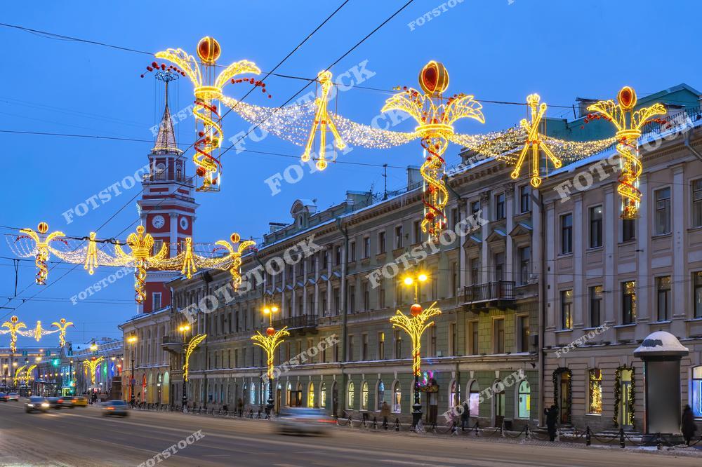 На фотографии изображен Невский проспект, украшенный новогодними огнями. Рождественская иллюминация в виде ангелов и ярких гирлянд создаёт атмосферу праздника. В центре внимания — башня, которая является важной архитектурной деталью на фоне вечернего города. Снежная улица и огни создают особую атмосферу зимнего Санкт-Петербурга в новогодние дни.